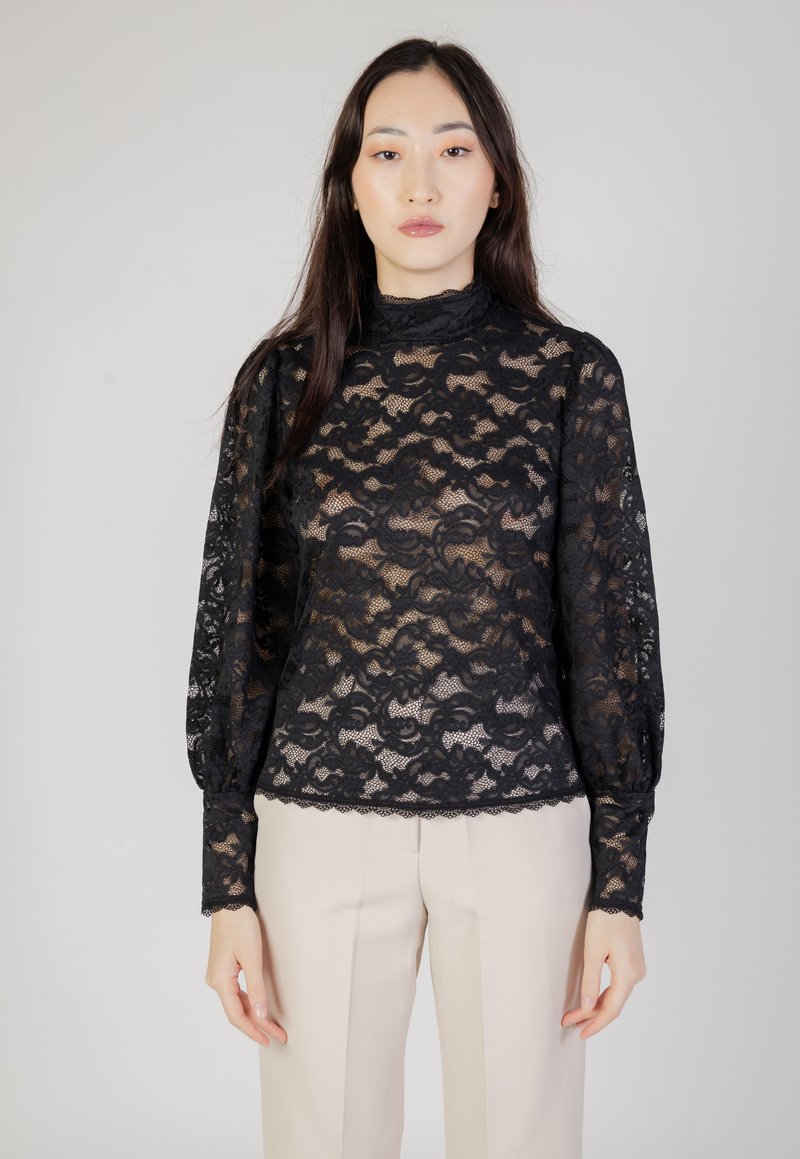 Schwarzes Spitzentop mit langen Puffärmeln, hohem Kragen und gewelltem Saum. Filigranes Blumenmuster; halbtransparente Textur. Kombiniert mit beigen Hosen.