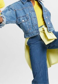 Zkrácená modrá džínová bunda se dvěma předními kapsami, pod níž je jasně žlutá košile. Outfit doplňují modré džínové kalhoty.