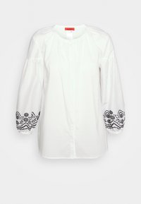 Blouse en coton blanc avec des manches longues bouffantes. Présente une broderie florale noire le long des poignets et un devant boutonné. Encolure ronde classique.