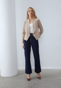 Beige avointa etureunaa oleva bleiseri, jossa on kolme neljäsosaa hihat, yhdistetty tummansinihin korkeavyötäröisiin suoriin farkkuihin ja ruskeisiin kuviollisiin korkokenkiin.