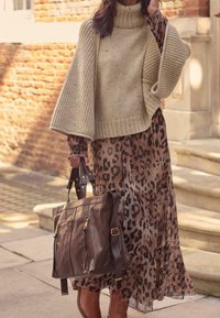 Femme portant un poncho en tricot beige et une robe à imprimé léopard, tenant un grand sac à main en cuir marron foncé, debout près de marches en pierre en plein air.