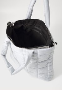Geantă de umăr quilted gri deschis cu fermoar, având un compartiment interior negru și două mânere rezistente. Textură fină și design căptușit.