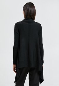 Mujer con el cabello corto y oscuro, vistiendo un largo cárdigan negro abierto con dobladillo asimétrico, combinado con pantalones negros, de pie de espaldas.