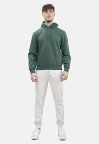Felpa verde con cappuccio dal taglio rilassato, polsini e vita a coste. Abbinata a pantaloni joggers grigi chiari a gamba stretta e sneakers bianche.