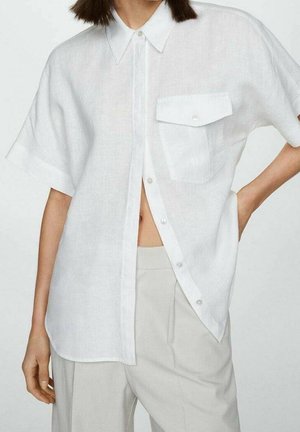 Femme portant une chemise blanche ample à manches courtes avec un bouton et une poche, associée à un pantalon taille haute beige clair, sur un fond uni.