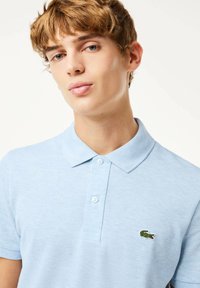 Ljusblå pikétröja gjord av texturerad bomull, med en tvåknappslucka och en broderad grön krokodil-logotyp på vänster bröst.