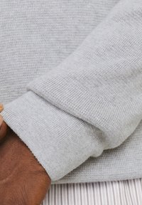 Lichtgrijze gebreide mouw van een sweater, gevouwen over een pols met donkere huid, die een fijne textuur en geribde manchetdetails toont.
