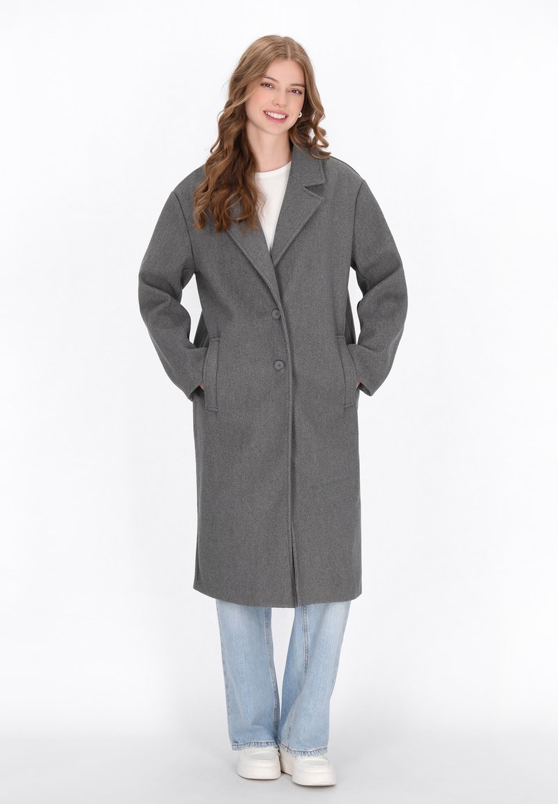 Cappotto in misto lana grigio con colletto a rever, chiusura frontale a due bottoni e tasche laterali. Indossato sopra una maglietta bianca e jeans azzurri chiari, abbinato a scarpe bianche.
