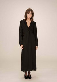 Robe noire à manches longues avec un décolleté en V profond, détail de devant twisté, tissu fluide et jupe longue, associée à des escarpins noirs pointus.