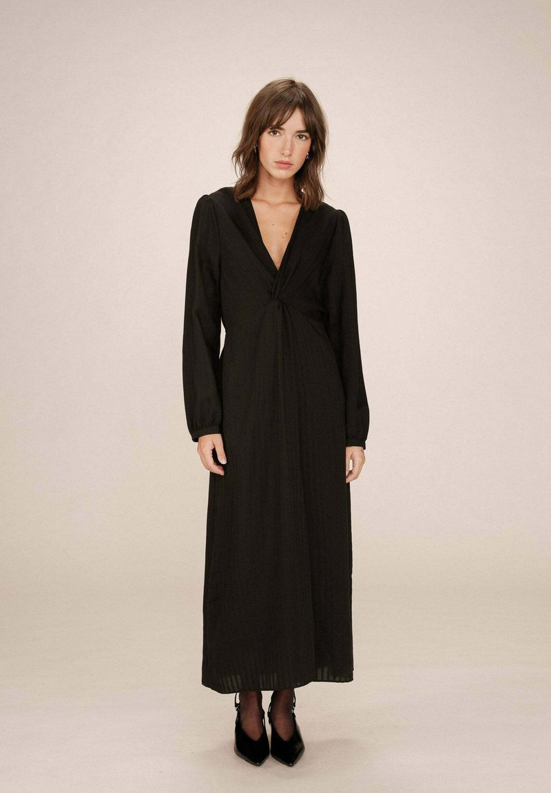 Robe noire à manches longues avec un décolleté en V profond, détail de devant twisté, tissu fluide et jupe longue, associée à des escarpins noirs pointus.