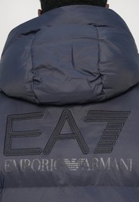 Oseba v temno modrem napihljivem jopiču z velikim oblazinjenim kapuco in logotipom "EA7 Emporio Armani" na hrbtu.