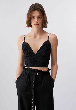 Młoda kobieta o krótkich brązowych włosach, ubrana w czarny haftowany krótki top na cienkich ramiączkach oraz wysokowalne czarne spodnie z paskiem.