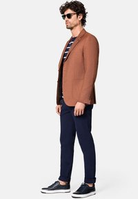 Brązowy żakiet o teksturze, granatowe spodnie, w paski koszula i ciemne sneakersy. Akcesoria to ciemne okulary przeciwsłoneczne i naszyjnik.