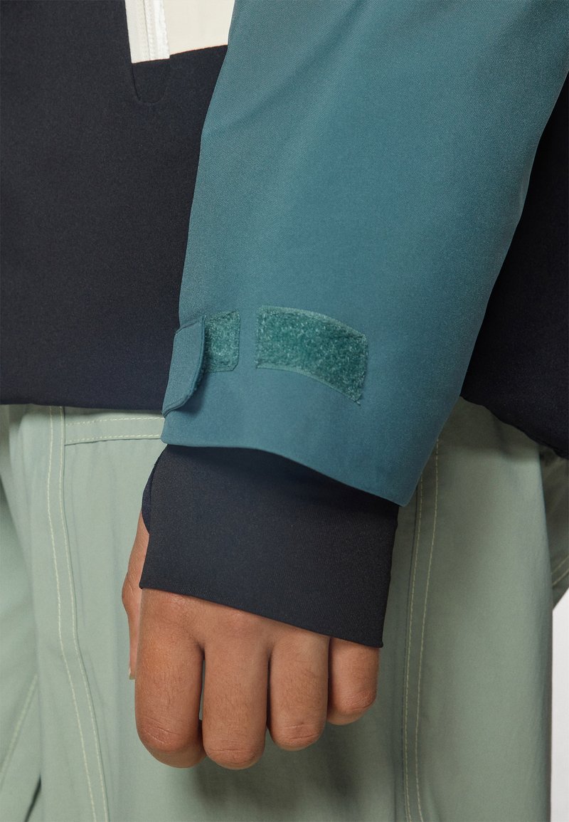 Ärmel einer türkis-schwarzen Jacke mit einem Klettverschluss, glatter Textur und einem eng anliegenden Bündchen, kombiniert mit hellgrünen Kleidungsstücken darunter.