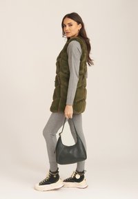 Olive green, plush puffer vest over a gray long-sleeve top, paired with gray leggings and chunky black sneakers. Holds a black handbag.