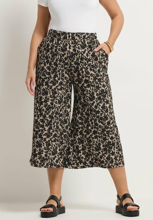 Kvinde iført vidde bukser i cropped længde med sort blomsterprint på beige, kombineret med sorte platformsandaler og en hvid top.