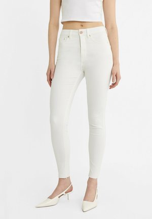 Femme portant un jean skinny taille haute blanc et des chaussures à talons slingback blanches, debout devant un fond blanc uni.