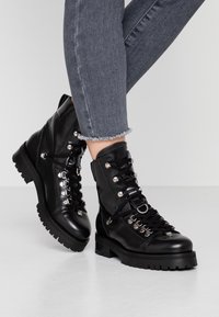 Bottes chevauchées en cuir noir avec des œillets métalliques, fermeture à lacets et semelles robustes. Présente une texture lisse et des accents en métal argenté.