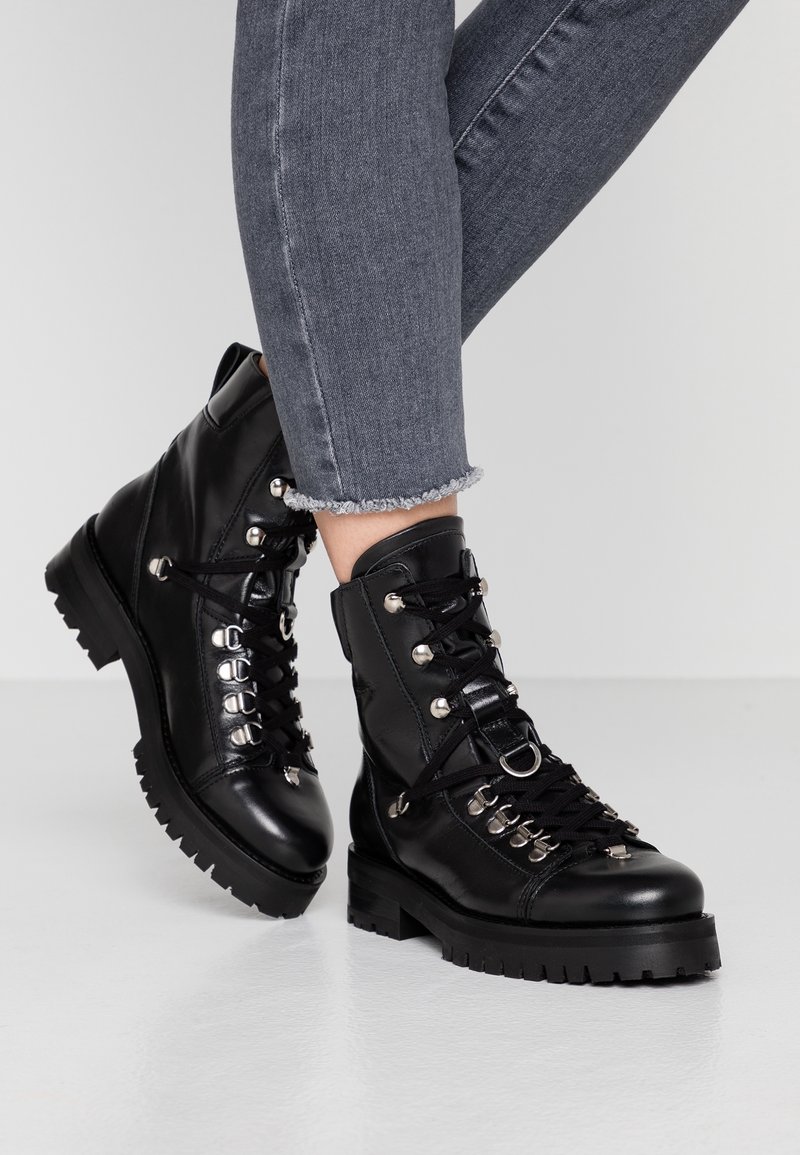 Bottes chevauchées en cuir noir avec des œillets métalliques, fermeture à lacets et semelles robustes. Présente une texture lisse et des accents en métal argenté.