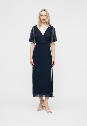 Kobieta w granatowej sukience kopertowej z przezroczystymi koronkowymi krótkimi rękawami i rozcięciem do uda, w białych sandałach na wysokim obcasie, stojąca na jednolitym tle.