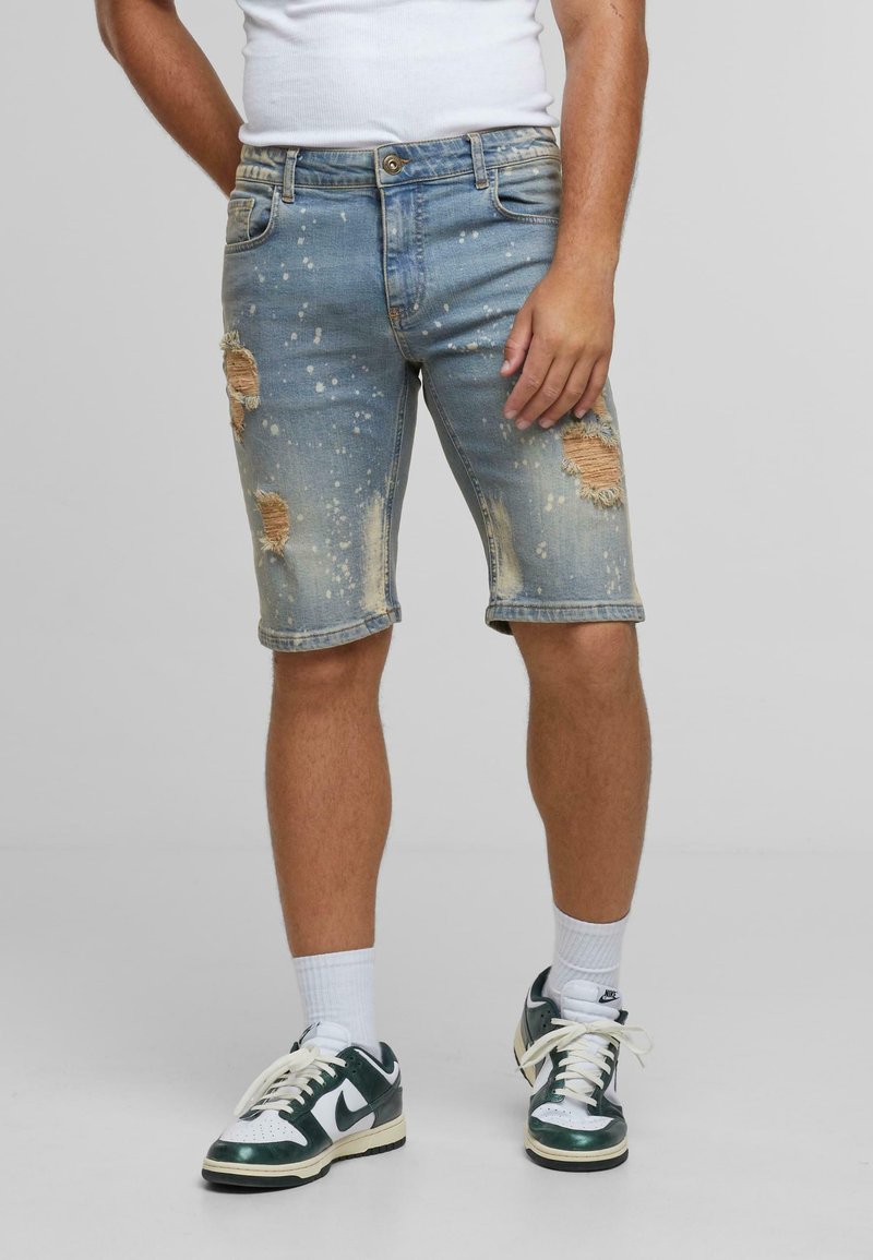 Traper šorts u svijetloj boji s isparanim mrljama i detaljima boje. Ima standardni pojas i rez do koljena.