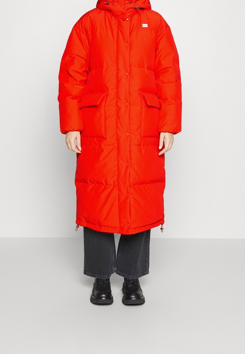Felrood pufferjack met een capuchon, drukknoopjes, twee voorzakken en een verstelbare zoom. Draag het over donkere, wijde jeans en zwarte schoenen.