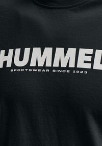 T-shirt en coton noir avec du texte blanc en bold "HUMMEL" et "SPORTSWEAR SINCE 1923." Col ras du cou classique et tissu texturé.