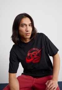 Juodas marškinėlis su raudonu grafiniu gyvatės ir skraidančio emblemos dizainu. Trumpomis rankovėmis ir apvaliu iškirpimu, pagamintas iš medvilnės mišinio.