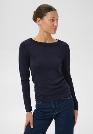 Une femme porte un pull bleu marine à manches longues et un jean bleu foncé sur un fond clair uni.