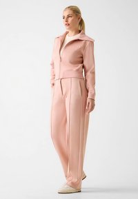 Veste zippée rose et pantalon large assorti en tissu doux. Dotés d’un col structuré et de passepoils contrastants le long des coutures.