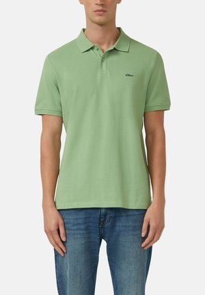 Man draagt een lichtgroene polo met korte mouwen, kraag en twee knopen, gecombineerd met een blauwe jeans tegen een effen achtergrond.