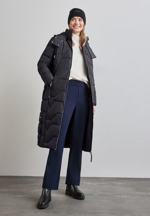 Manteau d'hiver noir avec un col montant, des poches zippées et une coupe droite. Porté par-dessus un léger pull et un pantalon bleu marine, avec des bottines noires.
