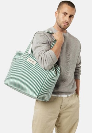 Homme portant un pull gris et un pantalon beige tenant un grand sac fourre-tout rayé vert et blanc sur l'épaule.