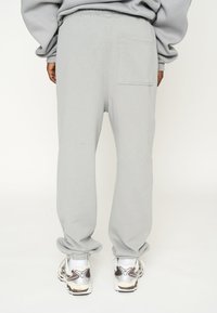 Pantalones de chándal gris claro con cintura elástica y un bolsillo trasero, con una textura suave y un ajuste relajado. Combinados con zapatillas blancas y grises.