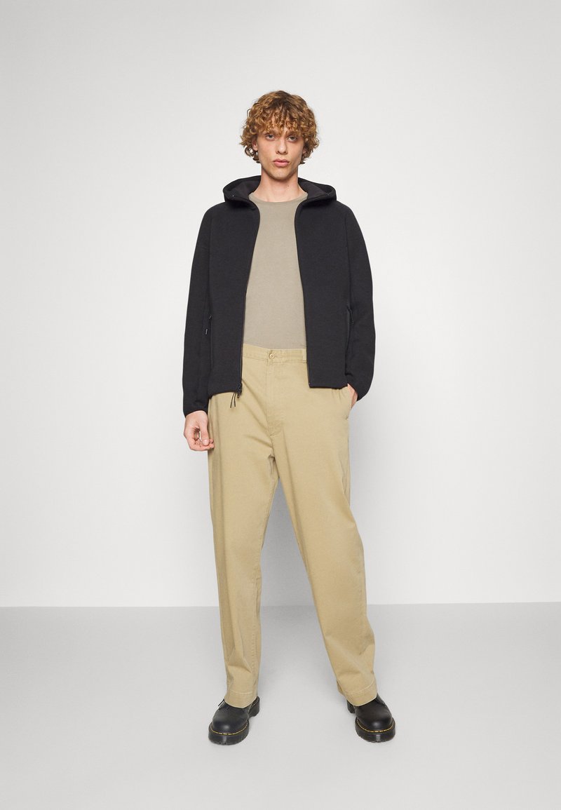 Svart zip-up hoodie, beige t-shirt, beige byxor, svarta skor. Hoodien har en huva, avslappnad passform och subtil textur. Enkelt och minimalistiskt design.