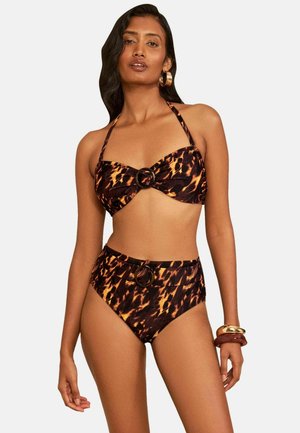Kobieta nosząca brązowo-czarny wzorzysty bikini halter z majtkami z wysokim stanem i okrągłymi klamrami, uzupełniony bransoletkami i kolczykami kołami.