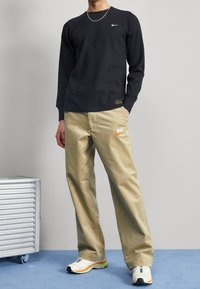 Chemise noire à manches longues en côte avec un logo Nike blanc, associée à un pantalon beige ample avec un logo Nike orange, et des chaussures de sport blanches.