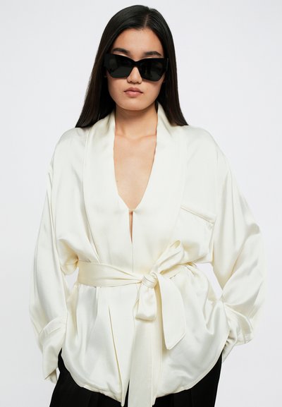 Λευκή σατέν μπλούζα με βαθύ V-λαιμόκοψη, φαρδύ γιακά και ζώνη στη μέση. Το μοντέλο φορά γωνιακά μαύρα γυαλιά ηλίου, προβάλλοντας την αντίθεση στο σχέδιο και το χρώμα.