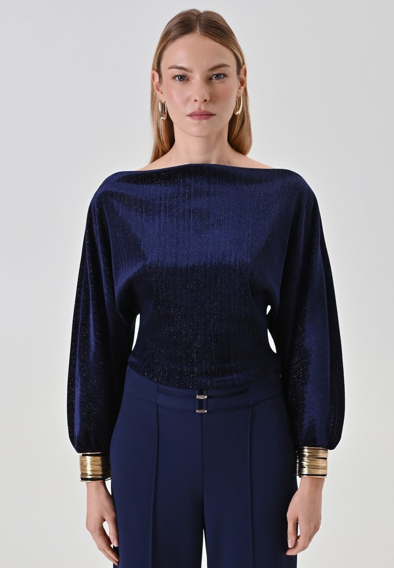 Blouse à manches longues bleu marine avec une texture brillante et un col off-épaule, présentant des poignets métalliques dorés et une coupe ajustée.