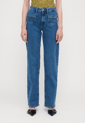 Frontalansicht einer Person, die hoch taillierte blaue Jeans mit großen Vordertaschen, ein olivgrünes, strukturiertes Oberteil und schwarze spitze Schuhe mit Schnallen trägt.