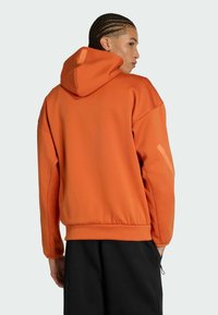 Felpa arancione con cappuccio, dal finish strutturato, spalle scese e un logo sul cappuccio. Abbinata a pantaloni neri larghi.