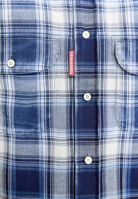 Modrá a biela kockovaná košeľa s dvoma náprsními vreckami, bielymi gombíkmi a červenou značkou Dsquared2 medzi vreckami.