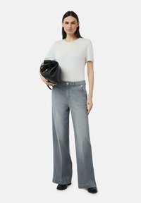 Weite graue Jeans mit vorderen Taschen und einem leichten, taillierten weißen T-Shirt. Das Model hält eine schwarze Bucket-Bag. Glatter, lässiger Look mit minimalen Details.
