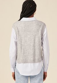 Gilet grigio lavorato a maglia con una morbida texture, design senza maniche e orlo a coste, indossato sopra una camicia a maniche lunghe blu chiaro e bianca con righe.