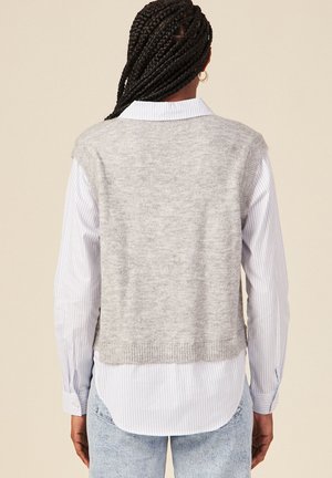 Grau gestrickte Weste mit weicher Textur, ärmellosem Design und geripptem Saum, über einem hellblauen und weißen gestreiften Langarmshirt getragen.