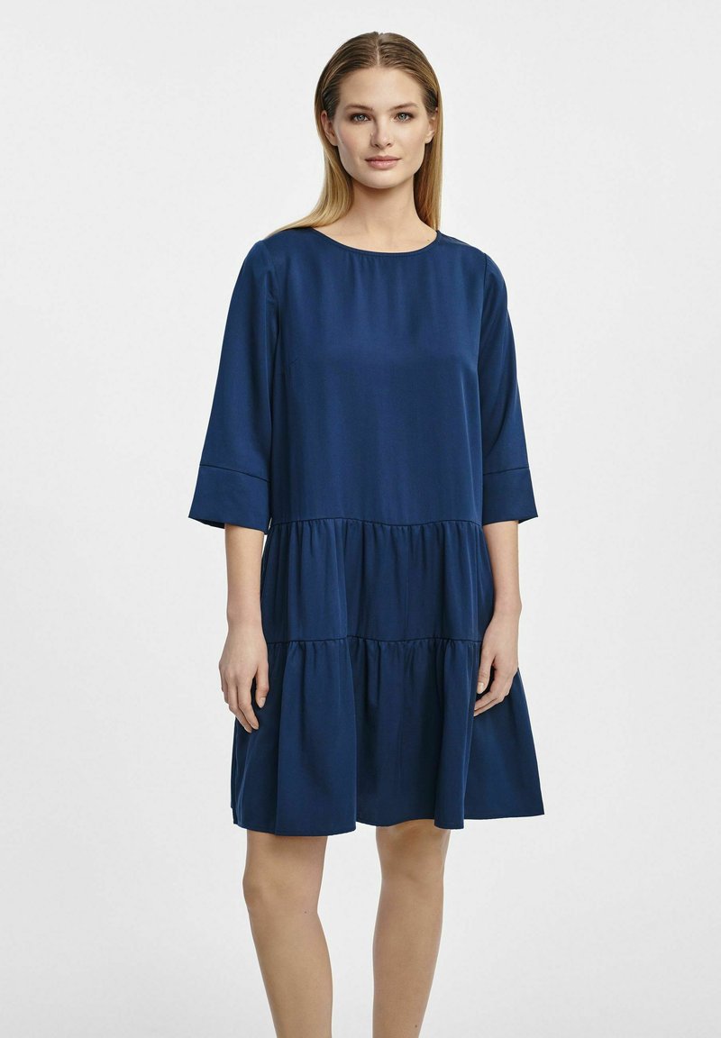 Granatowa sukienka z gładkiego materiału, z okrągłym dekoltem, rękawami o długości trzech czwartych oraz falbaniastym zdobieniem spódnicy.