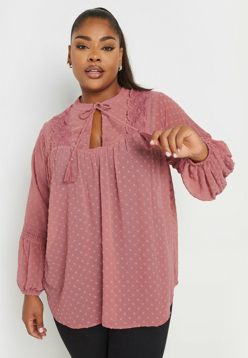 Blusa rosa confetto con pois testurizzati, scollatura a forma di chiave con fiocco, dettagli in pizzo e maniche a sbuffo voluminose. Tessuto morbido e fluente.