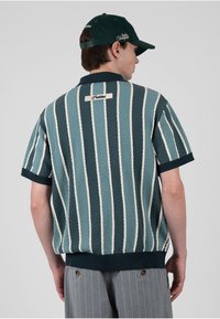 Hombre con camisa de manga corta a rayas verdes y blancas y gorra verde, de espaldas sobre un fondo gris liso.