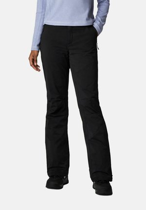 Personne portant un pantalon noir droit et des chaussures noires, avec une main dans la poche et un haut à manches longues bleu clair visible.