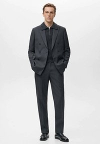 Abito doppiopetto grigio con camicia a maglia scura testurizzata e ampio colletto, abbinato a scarpe nere lucide, in piedi contro uno sfondo bianco.